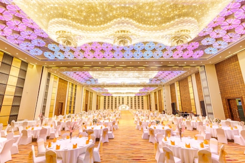 A large, elegantly decorated banquet hall with numerous round tables covered with white tablecloths and adorned with flowers. The ceiling features intricate lighting with patterns in shades of purple and gold. The floor appears to be a glossy wood finish under soft lighting, giving the room a luxurious feel.