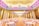 A large, elegantly decorated banquet hall with numerous round tables covered with white tablecloths and adorned with flowers. The ceiling features intricate lighting with patterns in shades of purple and gold. The floor appears to be a glossy wood finish under soft lighting, giving the room a luxurious feel.