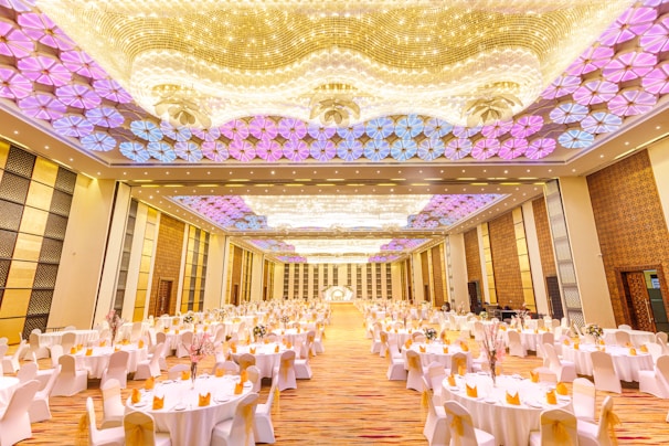A large, elegantly decorated banquet hall with numerous round tables covered with white tablecloths and adorned with flowers. The ceiling features intricate lighting with patterns in shades of purple and gold. The floor appears to be a glossy wood finish under soft lighting, giving the room a luxurious feel.