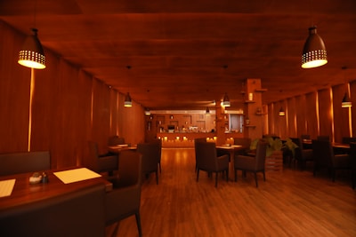 A warmly lit restaurant interior with wooden walls and flooring, featuring several dining tables and chairs. The lighting is provided by multiple hanging lamps with a cozy ambiance. The bar counter is visible at the far end with various bottles and glassware.