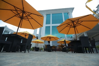 Large orange umbrellas are set up over outdoor dining tables with black wicker chairs on a paved patio. The background features a tall, modern building with large blue-tinted glass windows and white columns.