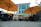 Large orange umbrellas are set up over outdoor dining tables with black wicker chairs on a paved patio. The background features a tall, modern building with large blue-tinted glass windows and white columns.