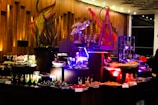 An elegant buffet setting with a variety of colorful desserts and drinks displayed on tables. Intricate sculptures, including a fish and a bird, provide decorative focal points. The ambient lighting with warm tones adds sophistication to the atmosphere.