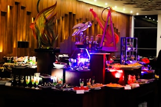 An elegant buffet setting with a variety of colorful desserts and drinks displayed on tables. Intricate sculptures, including a fish and a bird, provide decorative focal points. The ambient lighting with warm tones adds sophistication to the atmosphere.