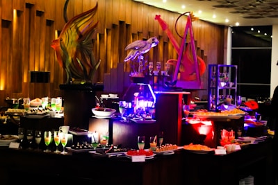 An elegant buffet setting with a variety of colorful desserts and drinks displayed on tables. Intricate sculptures, including a fish and a bird, provide decorative focal points. The ambient lighting with warm tones adds sophistication to the atmosphere.