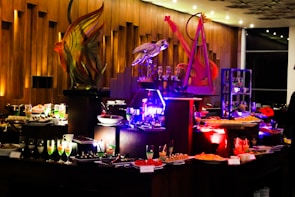 An elegant buffet setting with a variety of colorful desserts and drinks displayed on tables. Intricate sculptures, including a fish and a bird, provide decorative focal points. The ambient lighting with warm tones adds sophistication to the atmosphere.