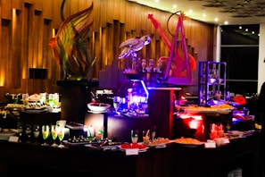 An elegant buffet setting with a variety of colorful desserts and drinks displayed on tables. Intricate sculptures, including a fish and a bird, provide decorative focal points. The ambient lighting with warm tones adds sophistication to the atmosphere.