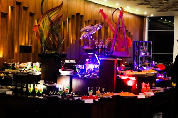 An elegant buffet setting with a variety of colorful desserts and drinks displayed on tables. Intricate sculptures, including a fish and a bird, provide decorative focal points. The ambient lighting with warm tones adds sophistication to the atmosphere.
