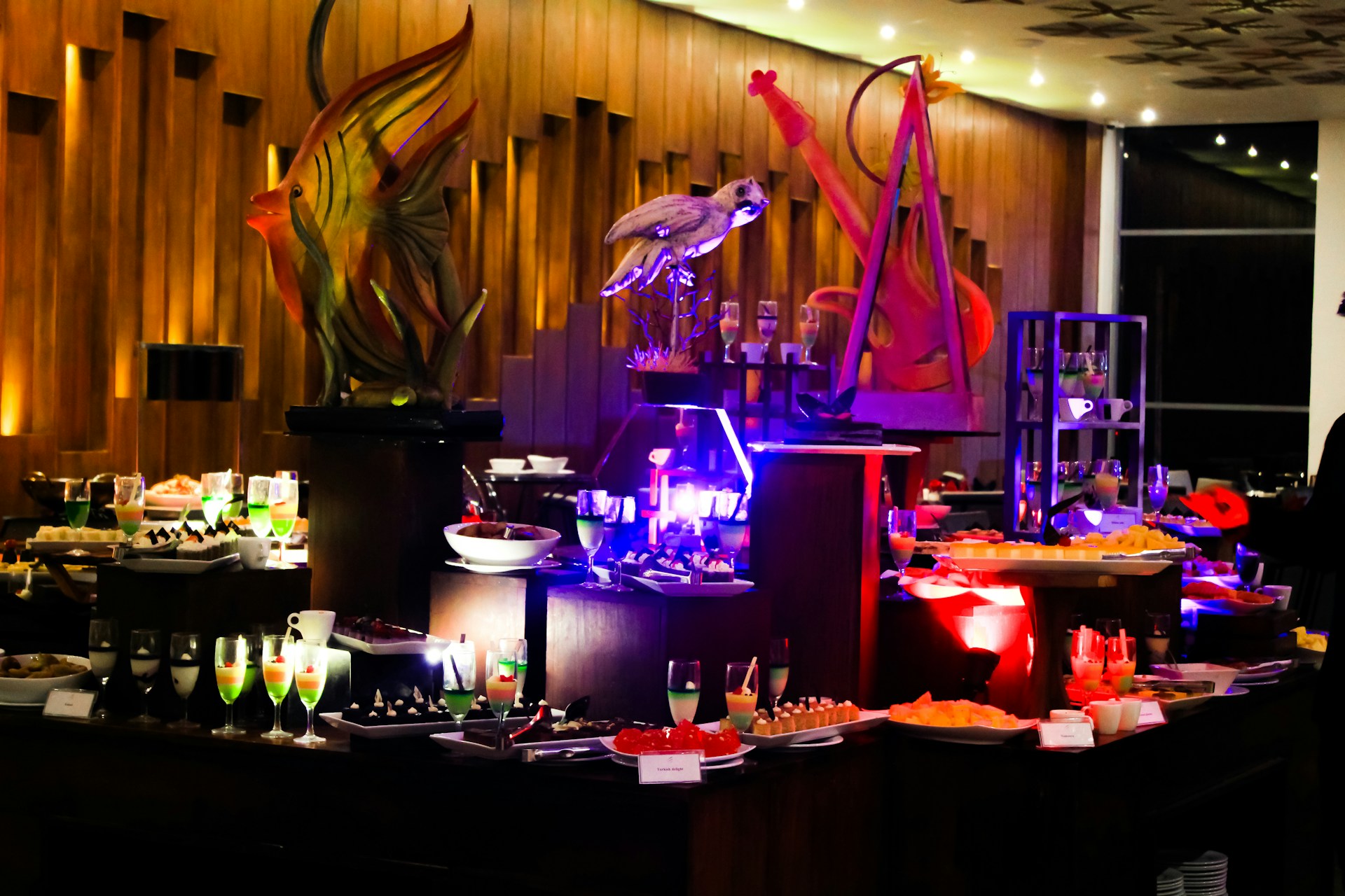 An elegant buffet setting with a variety of colorful desserts and drinks displayed on tables. Intricate sculptures, including a fish and a bird, provide decorative focal points. The ambient lighting with warm tones adds sophistication to the atmosphere.