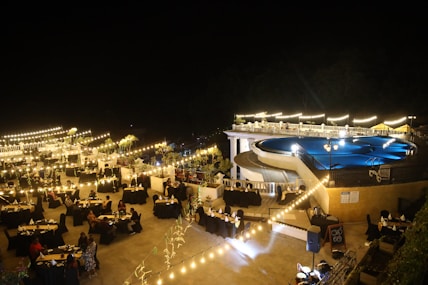 A nighttime outdoor event featuring a large pool surrounded by bright lights and elegantly arranged tables with black tablecloths. The ambiance is enhanced by string lights and decorative plants, creating a warm and inviting atmosphere. People are seated at the tables, suggesting a gathering or celebration.