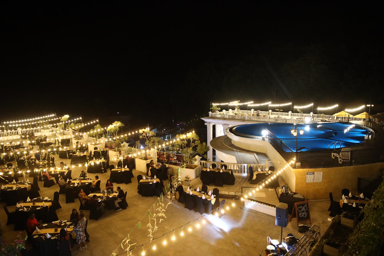 A nighttime outdoor event featuring a large pool surrounded by bright lights and elegantly arranged tables with black tablecloths. The ambiance is enhanced by string lights and decorative plants, creating a warm and inviting atmosphere. People are seated at the tables, suggesting a gathering or celebration.