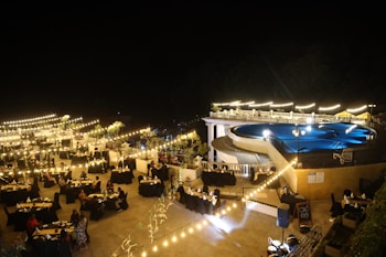 A nighttime outdoor event featuring a large pool surrounded by bright lights and elegantly arranged tables with black tablecloths. The ambiance is enhanced by string lights and decorative plants, creating a warm and inviting atmosphere. People are seated at the tables, suggesting a gathering or celebration.