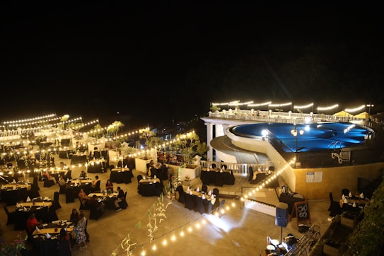 A nighttime outdoor event featuring a large pool surrounded by bright lights and elegantly arranged tables with black tablecloths. The ambiance is enhanced by string lights and decorative plants, creating a warm and inviting atmosphere. People are seated at the tables, suggesting a gathering or celebration.