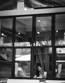 A black-and-white image captures two people sitting inside a cafe, visible through a large window. They appear to be in conversation, seated at a table with a vase containing tall decorative sticks. The setting is cozy, with soft lighting visible inside.