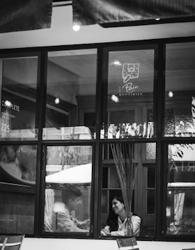 A black-and-white image captures two people sitting inside a cafe, visible through a large window. They appear to be in conversation, seated at a table with a vase containing tall decorative sticks. The setting is cozy, with soft lighting visible inside.