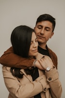 A young couple is embracing affectionately. The woman is wearing a beige coat and a wristwatch, while the man is in a brown jacket. They both have closed eyes, creating an intimate and serene atmosphere.
