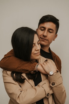 A young couple is embracing affectionately. The woman is wearing a beige coat and a wristwatch, while the man is in a brown jacket. They both have closed eyes, creating an intimate and serene atmosphere.