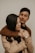 A young couple is embracing affectionately. The woman is wearing a beige coat and a wristwatch, while the man is in a brown jacket. They both have closed eyes, creating an intimate and serene atmosphere.