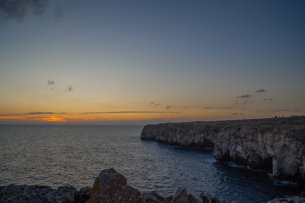 the sun is setting over the ocean near a rocky cliff