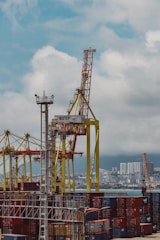 A bustling port filled with numerous colorful shipping containers stacked in layers. Large yellow cranes dominate the scene, towering over the containers, ready to load and unload them. In the distance, a city skyline is visible under a cloudy sky.