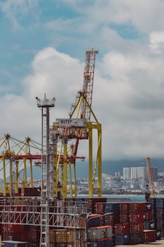 A bustling port filled with numerous colorful shipping containers stacked in layers. Large yellow cranes dominate the scene, towering over the containers, ready to load and unload them. In the distance, a city skyline is visible under a cloudy sky.