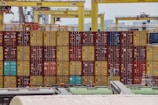 Rows of colorful shipping containers are stacked at a busy port. Numerous cranes labeled with 'Konecranes' are positioned above the containers. The containers exhibit a variety of colors including red, yellow, and blue. In the foreground, parts of industrial structures and equipment are visible.