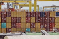 Rows of colorful shipping containers are stacked at a busy port. Numerous cranes labeled with 'Konecranes' are positioned above the containers. The containers exhibit a variety of colors including red, yellow, and blue. In the foreground, parts of industrial structures and equipment are visible.