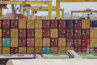 Rows of colorful shipping containers are stacked at a busy port. Numerous cranes labeled with 'Konecranes' are positioned above the containers. The containers exhibit a variety of colors including red, yellow, and blue. In the foreground, parts of industrial structures and equipment are visible.