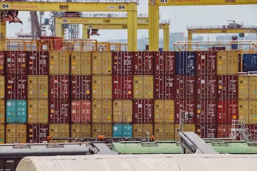 Rows of colorful shipping containers are stacked at a busy port. Numerous cranes labeled with 'Konecranes' are positioned above the containers. The containers exhibit a variety of colors including red, yellow, and blue. In the foreground, parts of industrial structures and equipment are visible.