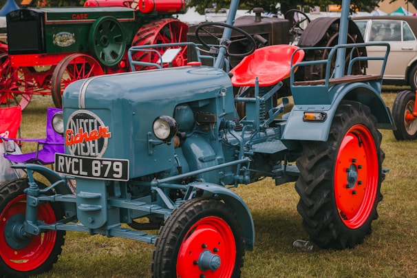 A vintage tractor is prominently displayed on a grass field. The tractor is painted in a blue color with vibrant red wheel rims. Other vintage machinery and vehicles are visible in the background, adding to a classic, nostalgic atmosphere. The tractor features a license plate and the brand 'Fischer' emblazoned on the front.