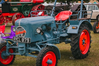 A vintage tractor is prominently displayed on a grass field. The tractor is painted in a blue color with vibrant red wheel rims. Other vintage machinery and vehicles are visible in the background, adding to a classic, nostalgic atmosphere. The tractor features a license plate and the brand 'Fischer' emblazoned on the front.