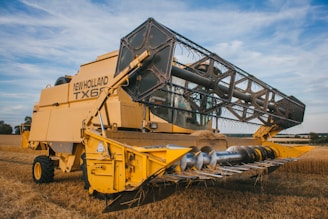 A large yellow combine harvester is parked in a field of wheat. The machine is adorned with the brand name 'New Holland TX66' and appears well-suited for harvesting crops. The background features a partly cloudy sky and additional fields extending into the distance.