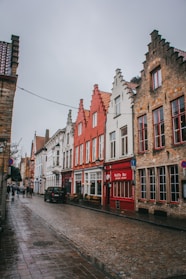 A quaint European street lined with colorful, traditional brick buildings featuring stepped gables. The cobblestone road is wet, suggesting recent rain, and a few people can be seen walking in the distance. A prominent red building houses a waffle bar.