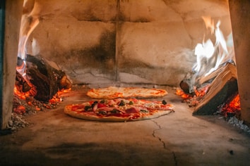 Two pizzas are being cooked inside a traditional wood-fired oven. Flames and embers surround the pizzas, creating a warm and rustic atmosphere. The oven is made of stone, and the pizzas are topped with various ingredients such as pepperoni and vegetables.