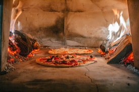 Two pizzas are being cooked inside a traditional wood-fired oven. Flames and embers surround the pizzas, creating a warm and rustic atmosphere. The oven is made of stone, and the pizzas are topped with various ingredients such as pepperoni and vegetables.