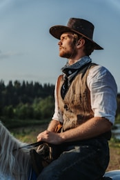 A man dressed in traditional Western attire, including a brown cowboy hat, bandana, and vest, is riding a horse. He gazes thoughtfully into the distance with a backdrop of lush, tree-filled landscape under a clear blue sky.