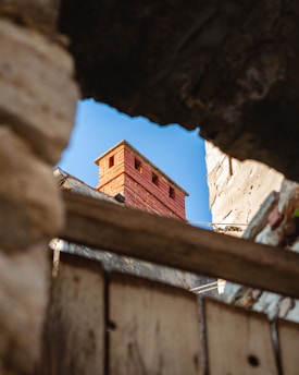 A weathered red brick chimney rises above a rooftop, surrounded by a clear blue sky. It is framed by rough, broken edges of a wall or structure, suggesting an old or abandoned setting.