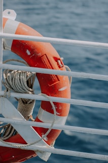 A bright orange lifebuoy with white accents is secured to the railing of a boat. Thick rope is wrapped around the lifebuoy, and the railing is painted white. The background features a body of water with gentle ripples, indicating a calm sea.