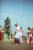 A group of women dressed in formal attire, walking down a street. One woman in a white dress and hat holds a bouquet of flowers, while others in pastel dresses are engaged with each other. The street is lined with houses and trees, under a clear blue sky.