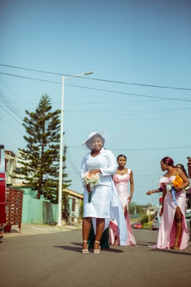 A group of women dressed in formal attire, walking down a street. One woman in a white dress and hat holds a bouquet of flowers, while others in pastel dresses are engaged with each other. The street is lined with houses and trees, under a clear blue sky.