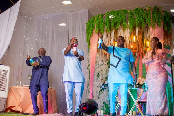 Four people stand in a celebratory setting, where three men are opening bottles of champagne with sparkling liquid spraying out. They are dressed in traditional and formal attire, standing in front of curtains and green foliage with decorative lights. A woman stands to the side, clapping her hands.