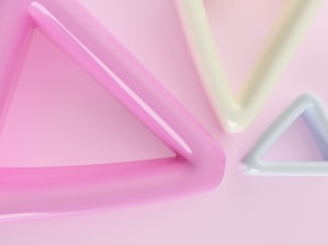 Three large, glossy triangles of varying sizes and colors are positioned against a soft pink background. The triangles have a smooth, reflective surface, with the colors being pink, pale yellow, and light blue, creating a harmonious and gentle visual effect.