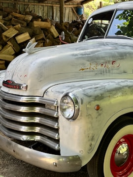 The image depicts the front portion of a vintage Chevrolet truck with its distinctive chrome grille and headlights. The truck's body has a slightly weathered appearance with faded paint and visible wear over time. In the background, there is a stack of chopped wood under a rustic wooden shelter.