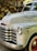 The image depicts the front portion of a vintage Chevrolet truck with its distinctive chrome grille and headlights. The truck's body has a slightly weathered appearance with faded paint and visible wear over time. In the background, there is a stack of chopped wood under a rustic wooden shelter.