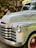 The image depicts the front portion of a vintage Chevrolet truck with its distinctive chrome grille and headlights. The truck's body has a slightly weathered appearance with faded paint and visible wear over time. In the background, there is a stack of chopped wood under a rustic wooden shelter.