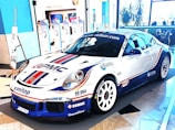 A sleek, white sports car decorated with various sponsor logos on its body. It's displayed in an indoor setting with a modern almost minimalist interior, including blue and white walls, large windows, and promotional banners in the background.