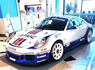 A sleek, white sports car decorated with various sponsor logos on its body. It's displayed in an indoor setting with a modern almost minimalist interior, including blue and white walls, large windows, and promotional banners in the background.