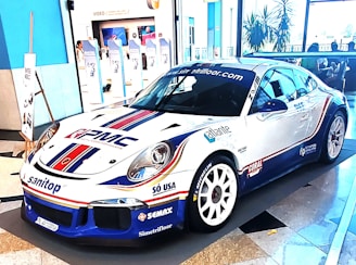 A sleek, white sports car decorated with various sponsor logos on its body. It's displayed in an indoor setting with a modern almost minimalist interior, including blue and white walls, large windows, and promotional banners in the background.