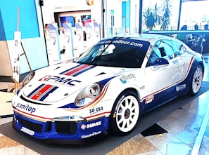 A sleek, white sports car decorated with various sponsor logos on its body. It's displayed in an indoor setting with a modern almost minimalist interior, including blue and white walls, large windows, and promotional banners in the background.
