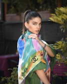 A woman with a high ponytail is wrapped in a colorful Chanel scarf featuring a variety of vibrant colors and patterns. She is standing amidst some greenery, with a confident expression and wearing a stylish watch. The background appears to be blurred, highlighting her as the main subject.